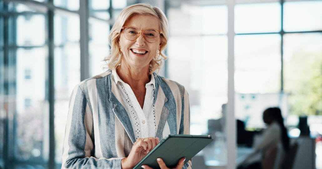 Professora idosa sorridente segurando um tablet em uma sala de aula moderna e bem iluminada, transmitindo educação de qualidade e tecnologia na educação.