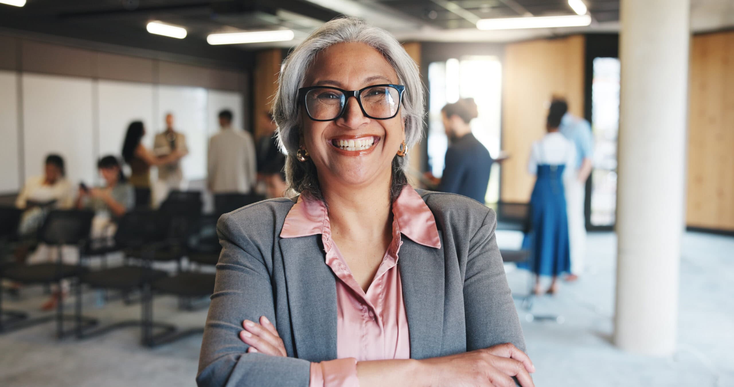 Professora sorridente com óculos, vestindo blazer cinza e blusa rosa, em ambiente de escola com estudantes ao fundo.