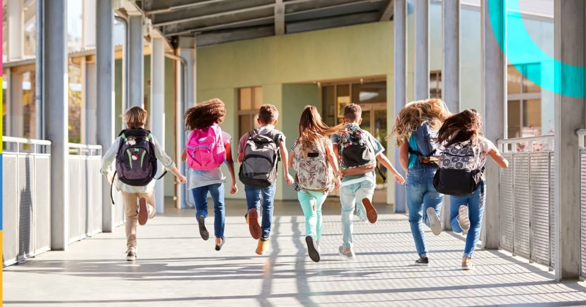 Grupo de crianças com mochilas caminhando em direção à escola em um ambiente externo.