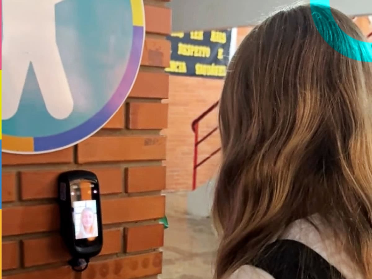 Estudante com cabelo castanho claro usando dispositivo eletrônico de reconhecimento facial para a chamada escolar automática na escola, com parede de tijolos ao fundo e marca do supersistema Diário Escola. Tecnologia nas redes municipais de ensino em uma escola pública de Planalto (RS)